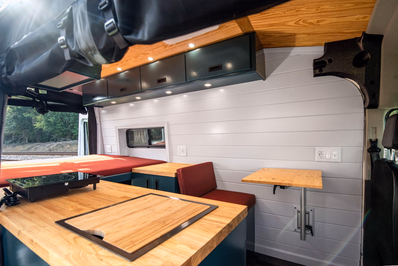Custom camper van kitchen with natural wood countertops, overhead cabinets, an induction cooktop, and a dinette.