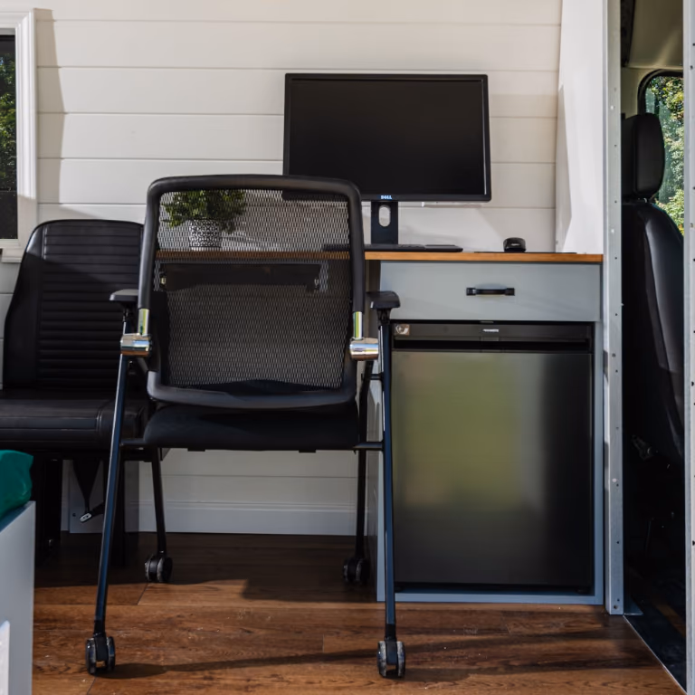Computer workstation inside the van with a monitor, desk chair, wood countertop, drawer, and under-counter refrigerator.