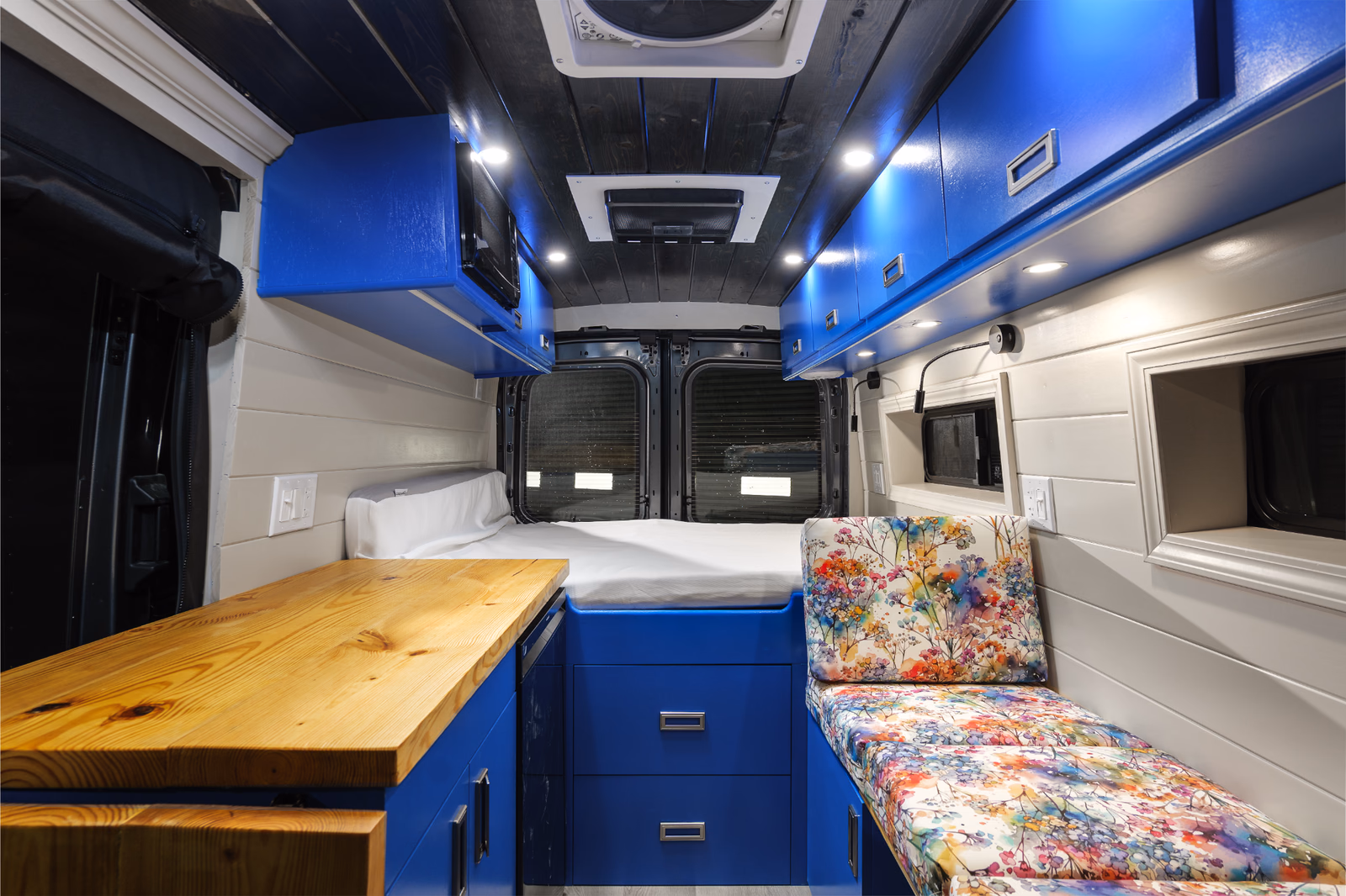 Camper van interior with blue painted cabinets, wood countertop, floral upholstery, and white ceiling lights.